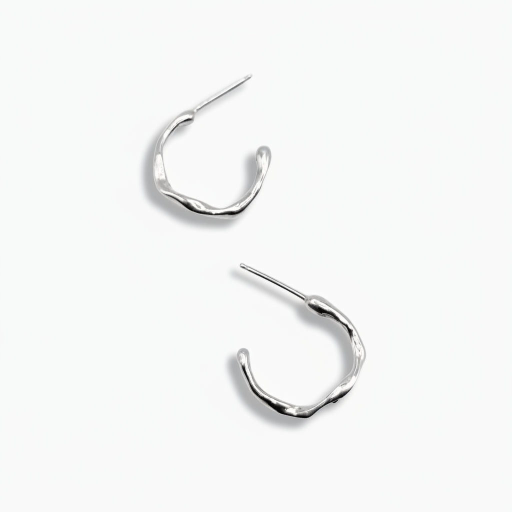 A pair of small, silver, hoop earrings with a wavy, organic surface on a white background.