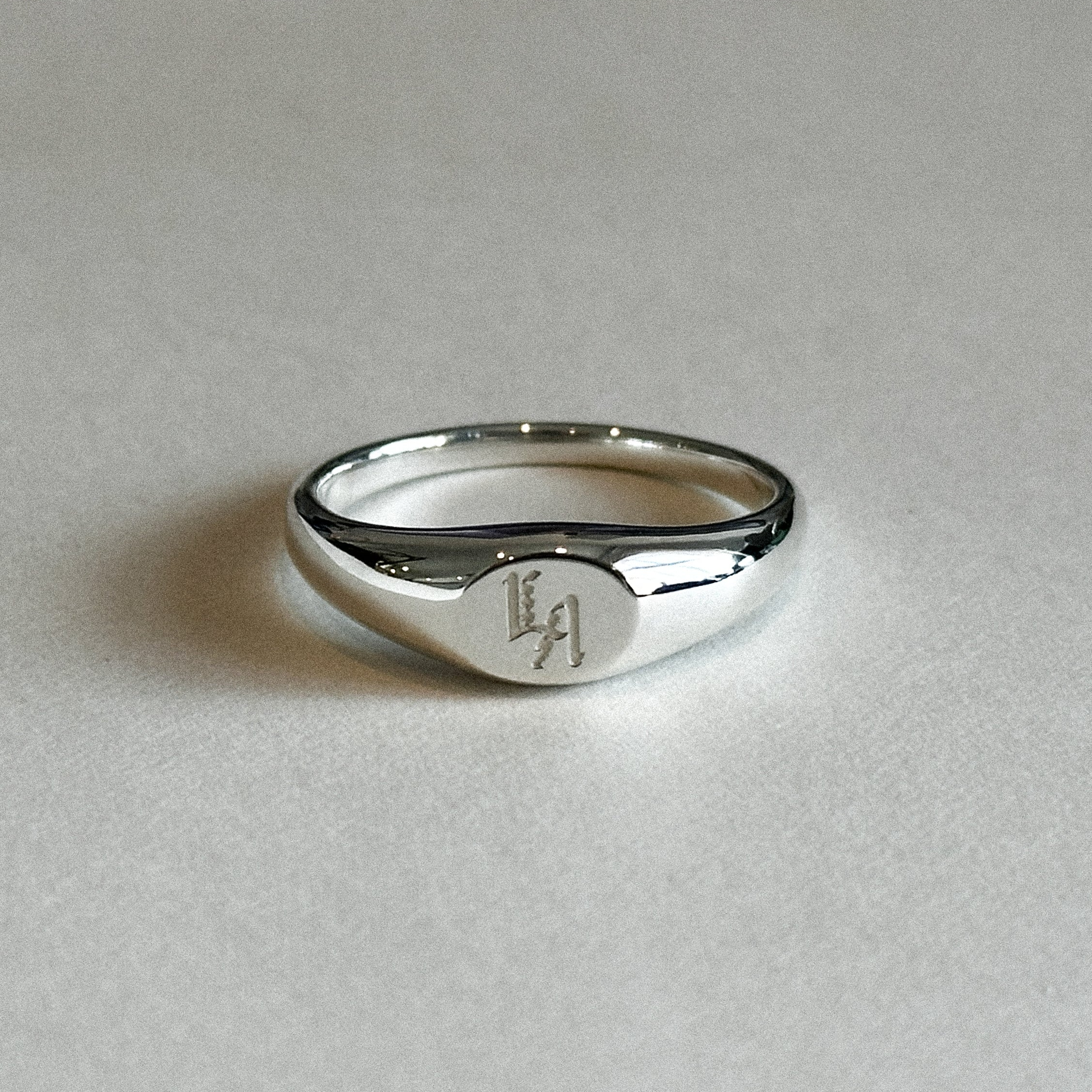 Silver signet ring with an AK ID LA mark engraving on a light gray background