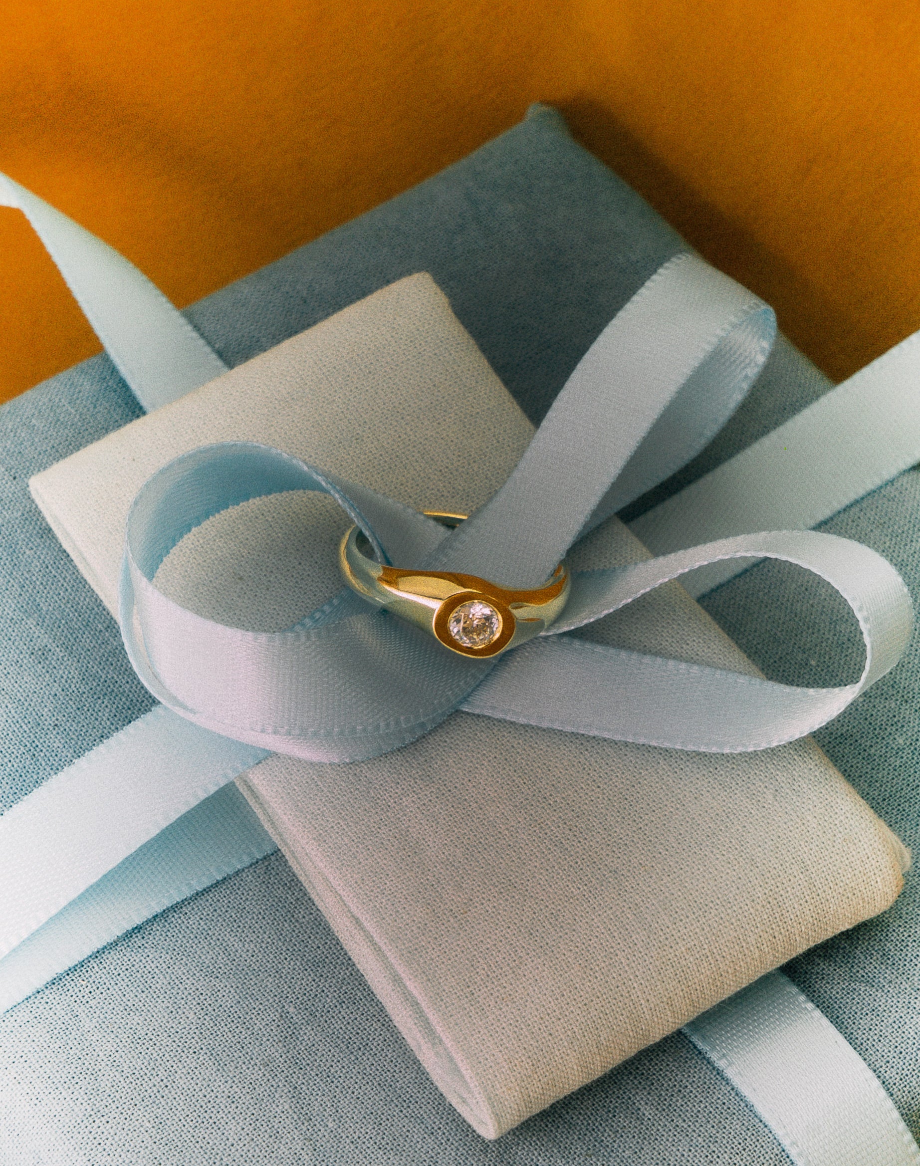 Gold ring with a diamond on a light blue ribbon over a beige box against an orange background